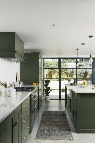 Green light shaker kitchen with Crittal-style windows, with green cabinets and white marble countertops, a large island to the right, and a small grey rug on the white floor, and hanging lights over the island