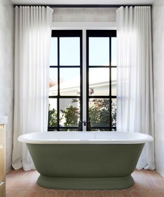 Large crittall framed windows with white drapes behind a green bath tub.