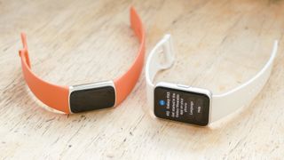 Close-up of the Samsung Galaxy Fit3 fitness tracker on a white rubber strap next to a Fitbit Charge 6 on an orange strap; both are sitting on a wood table