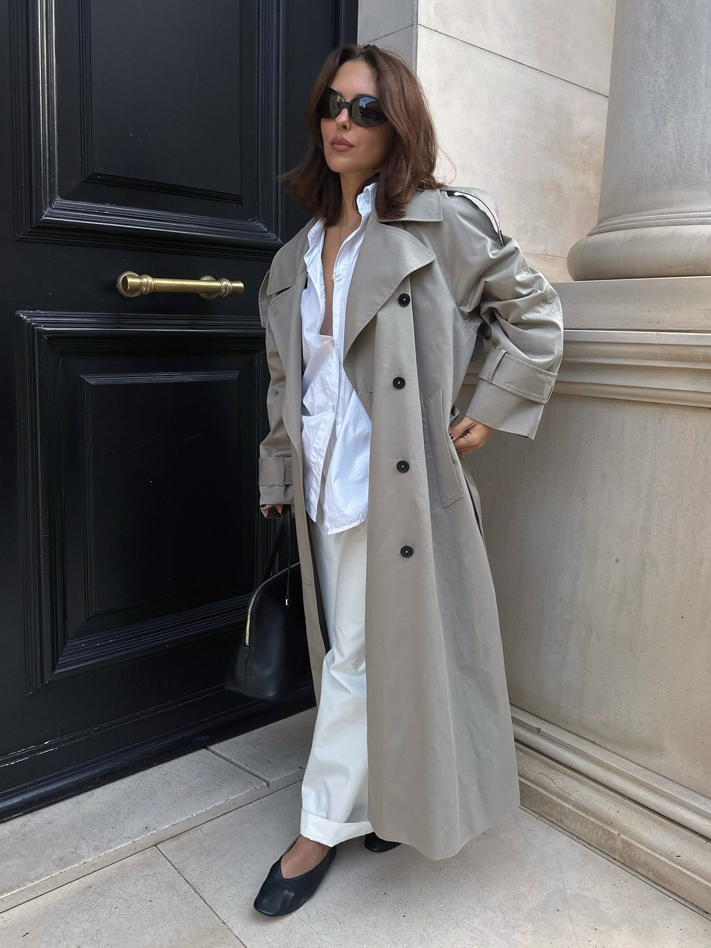 Style influencer Deborah Rosa wearing a long trench coat, white button-down shirt, cuffed white pants and black V-cut ballet flats