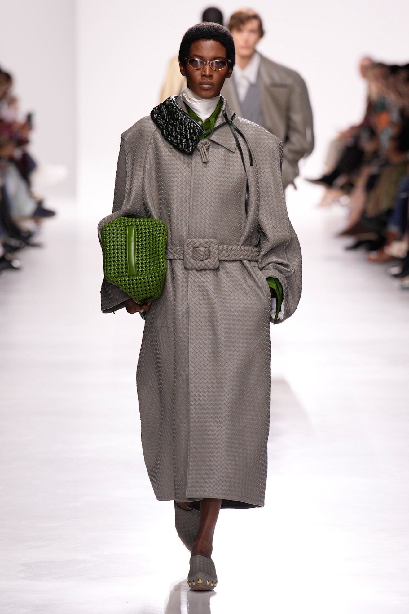 A model walking in the S/S 26 Bottega Veneta show wearing a gray Intrecciato trench coat.
