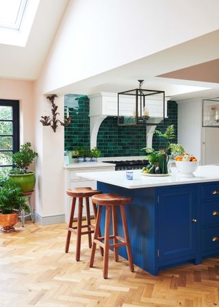 A kitchen by Neptune with blue and white shaker-style island, two wooden bar stools, green tiles around hob area and wood effect flooring