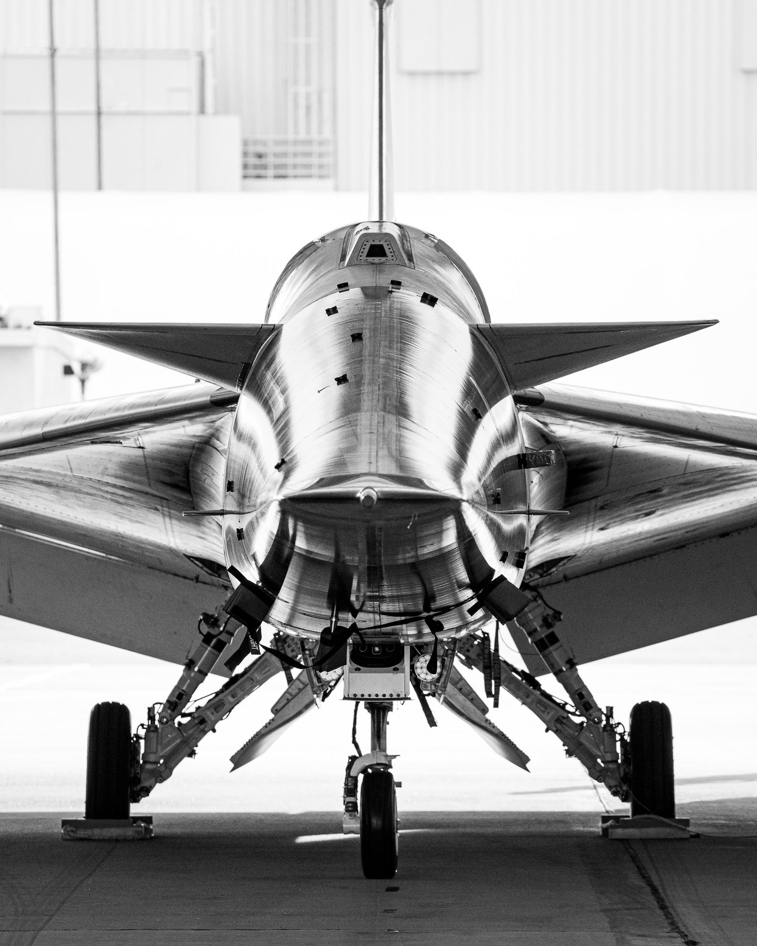 A head-on black and white image of the X-59 supersonic jet. The jet sits on three-wheeled landing gear, and the sharp, sleek angles of the jet's body and wings reflect as shapely light, highlighting the craft's curves and details.