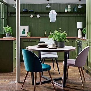 An olive green kitchen with green walls, dining table in the foreground and pastel pink and teal upholstered dining chairs.