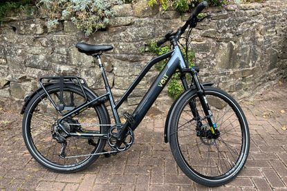This is an image of the Volt Pulse LS hybrid e-bike on a brick paved drive with a stone wall and greenery in the background