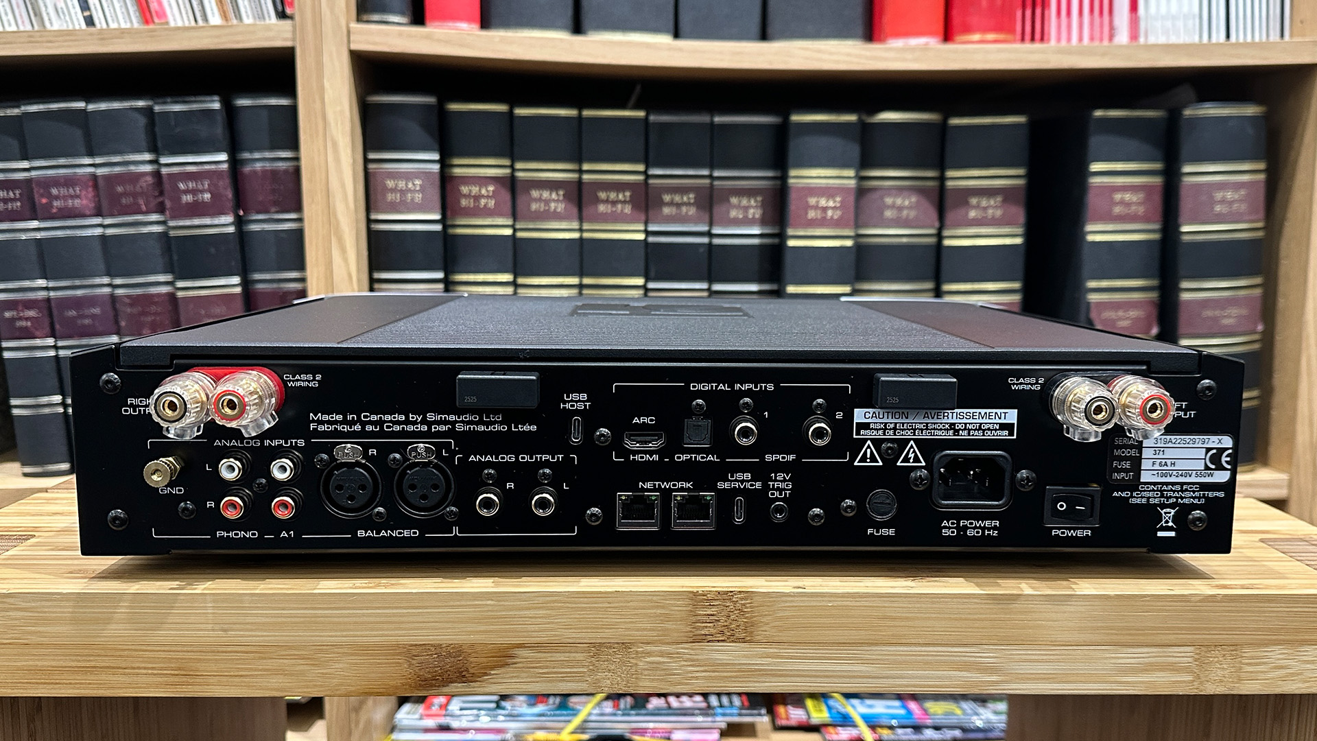 Moon 371 streaming amplifier on wooden hi-fi rack in front of bookcase, rear of unit showing connections
