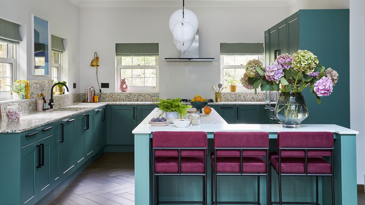 Teal kitchen with terrazzo worktops, dark wood parquet flooring, white globe pendant lights and bright pink bar stools