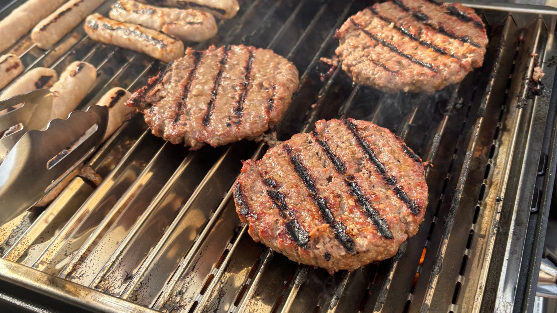 Burgers and sausages sizzling on a Char-Broil Grill2Go X200 portable gas barbecue, with visible chargrill marks as they cook during a garden barbecue.