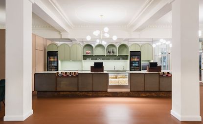Interior view of the bar area at Yorck Kino Passage cinema featuring brown floors, white pillars, a brown bar counter with a section for popcorn, sphere chandeliers, black fridges, pastel pink units and arched pastel green units with shelving