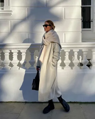 Rosie Huntington-Whiteley wears a white long coat with a white sweater tied around her shoulders, black sunglasses, and The Row's zip-up boots in black.