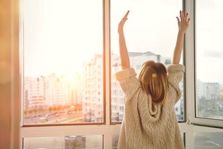 A woman streches in front of a large window in the morning sun.