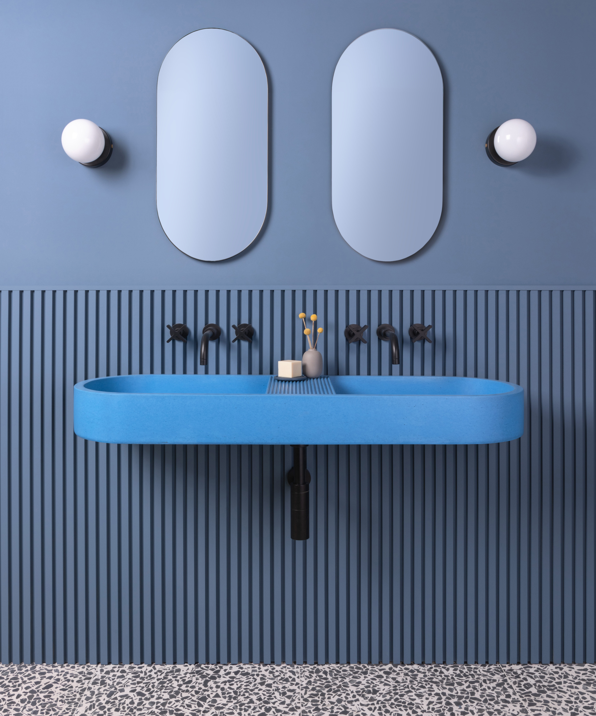 bathroom with blue terrazzo floor, ribbed blue wall panelling, modern blue wall mounted double sink, 2 oval mirrors and two white glass globe wall lights