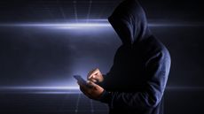 A man in a dark hoodie with the hood up uses a smartphone against a dark background.