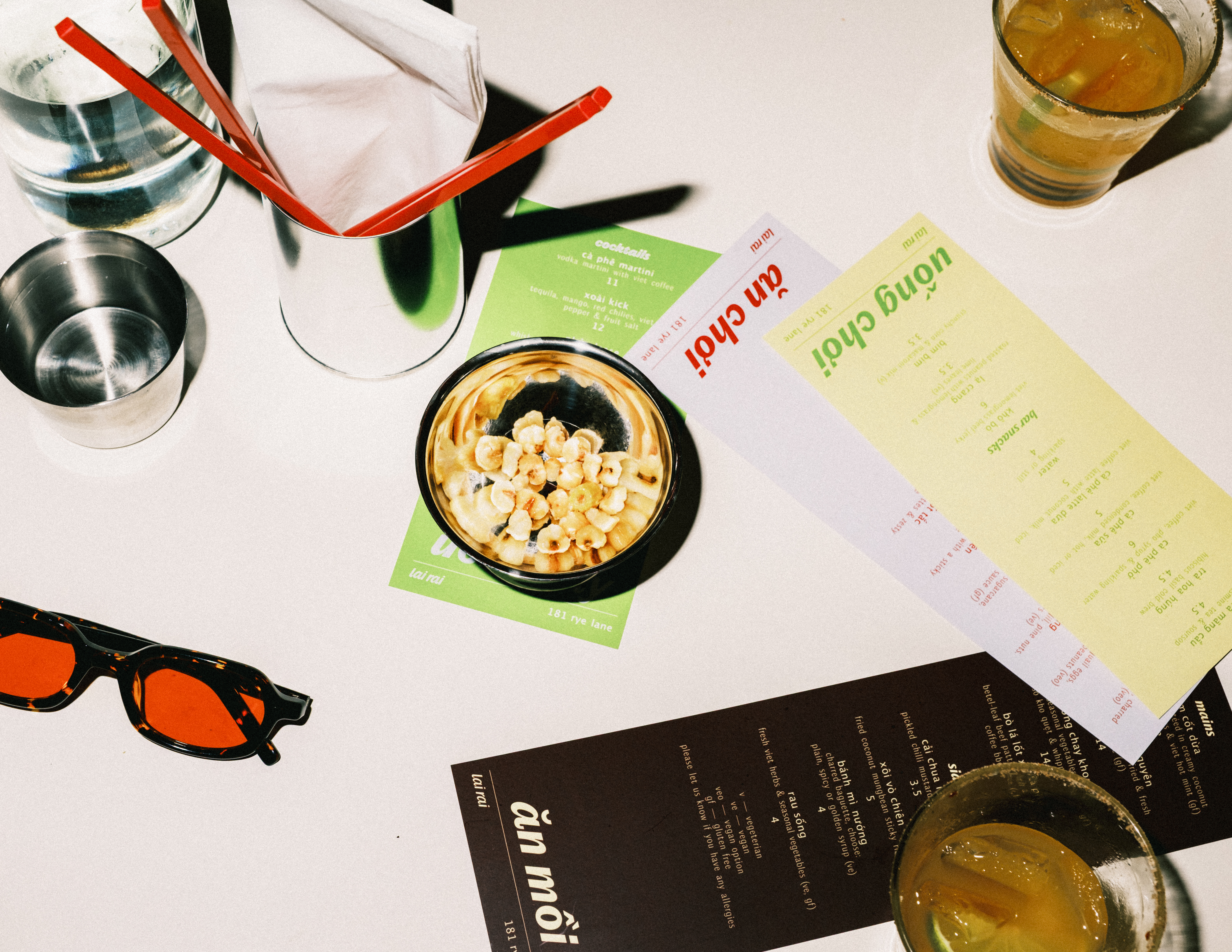 A set of colorful menus sits on a white table along with chrome cups, red chopsticks, a pair of black and red sunglasses, and drinks.