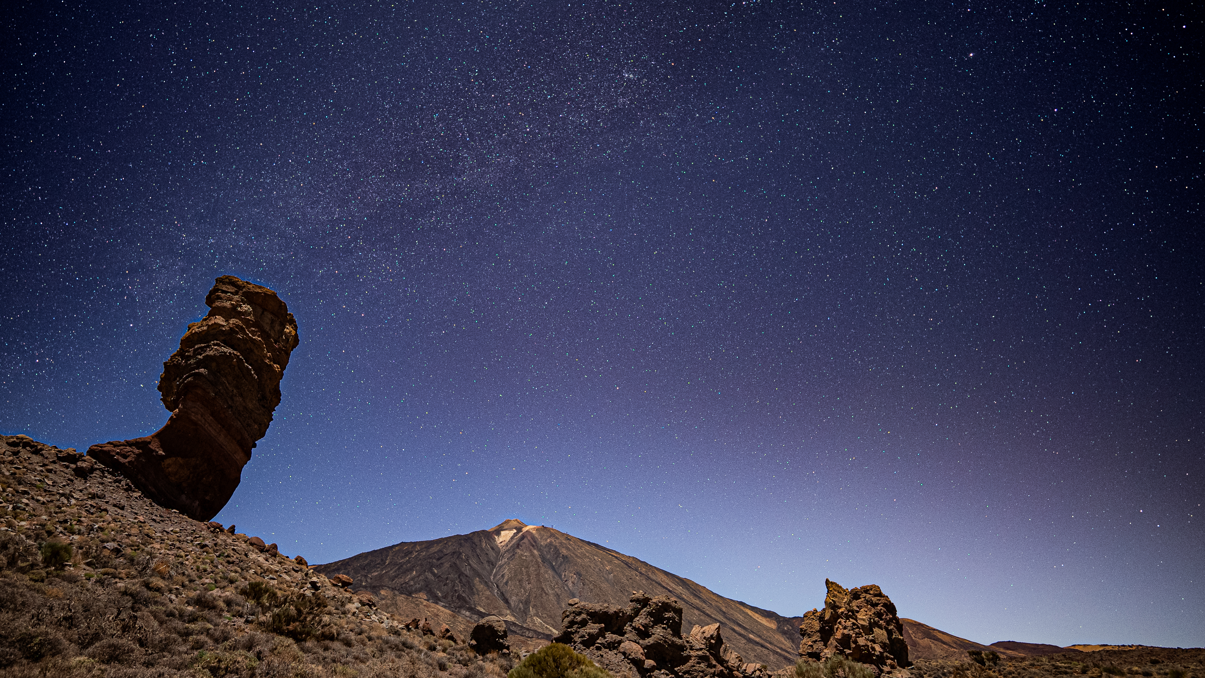 rock formations in the foreground, a starry night sky in the background and a distant volcano.
