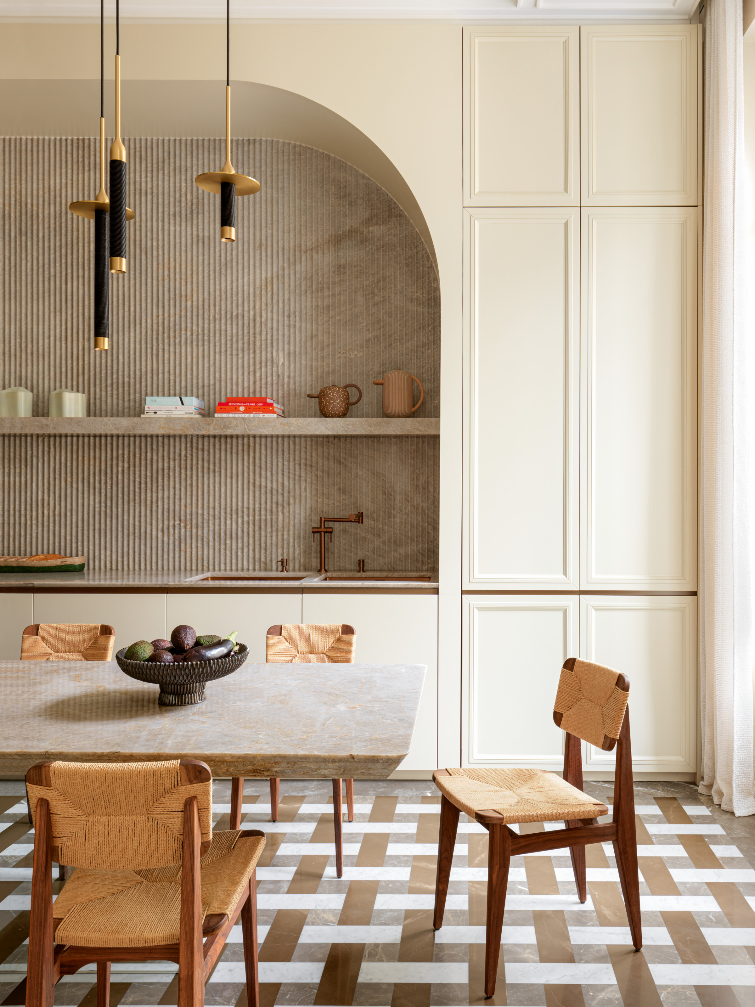 Cream kitchen with arched joinery, fluted splashback, geometric grey and bronze floor, stone dining table and woven chairs
