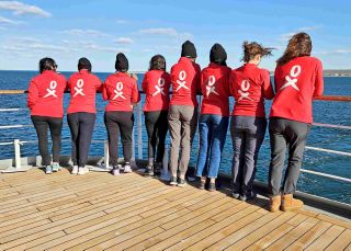 Women in matching red shirts stand on a ship, looking out at the sea. Their backs are facing the camera.