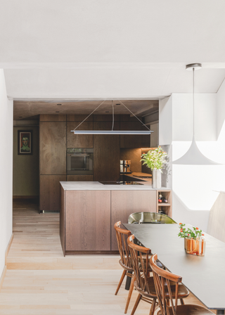 A pale dining area leading to a dark, wood cladded kitchen