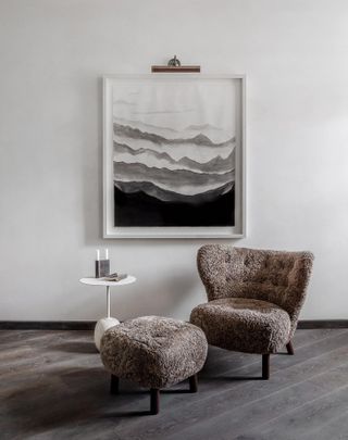 A sitting area with a grey fluffy chair and foot stool, a round coffee table below a large wall painting.