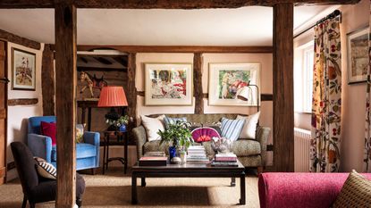 A cozy, rustic living room with exposed dark wood beams and pale pink walls. A leopard-print sofa with patterned cushions is centered around a dark wood coffee table stacked with books. Two framed artworks hang above the sofa. A bright red lamp and blue armchairs add pops of color