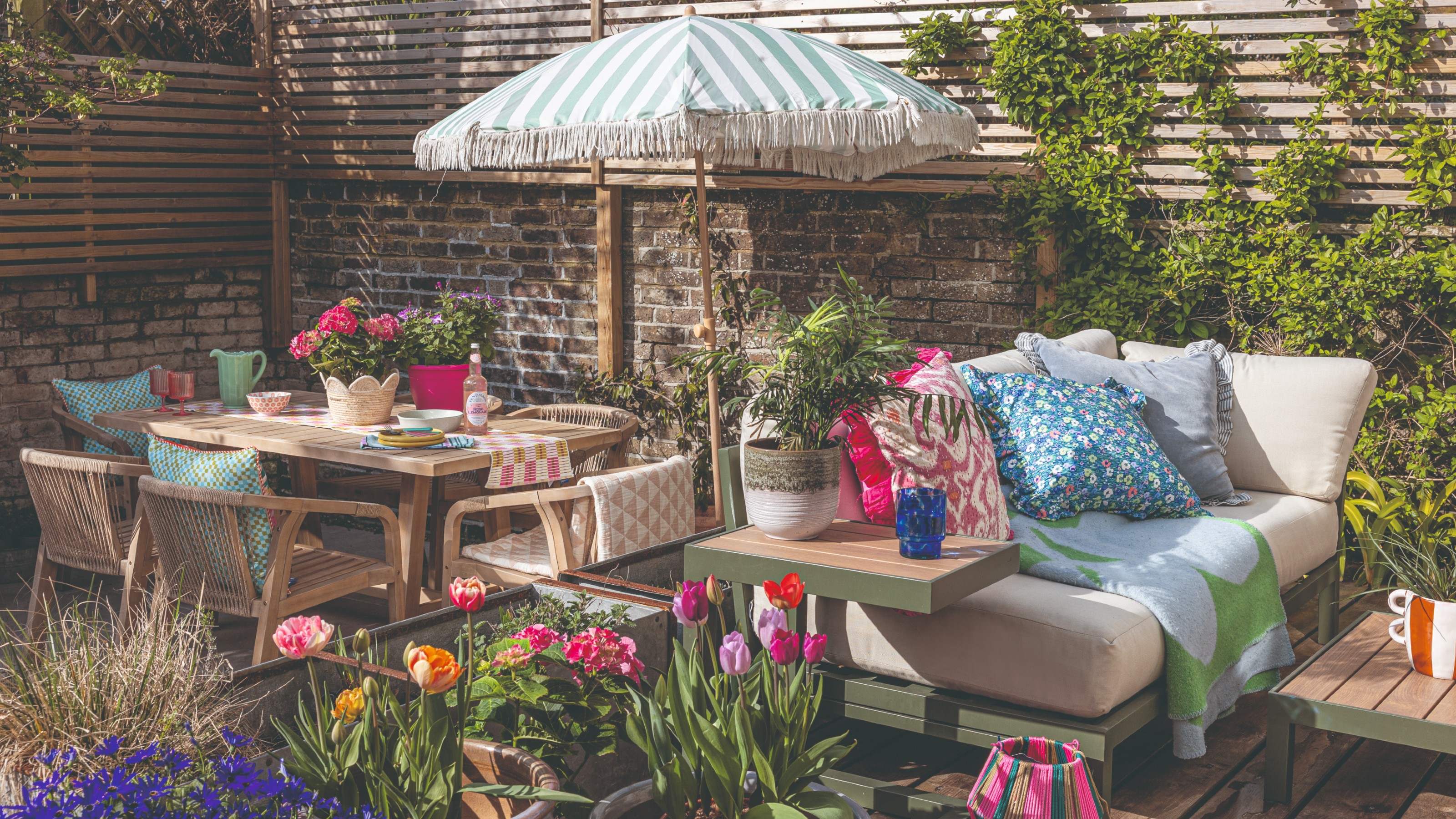 A garden with an outdoor sofa adorned with colourful outdoor cushions set next to a dining area with a striped fringed parasol