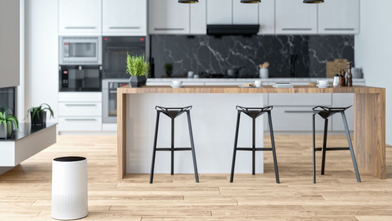 A white air purifier in a modern wooden and white kitchen, with black bar stools, black pendant lights and black marble backsplash