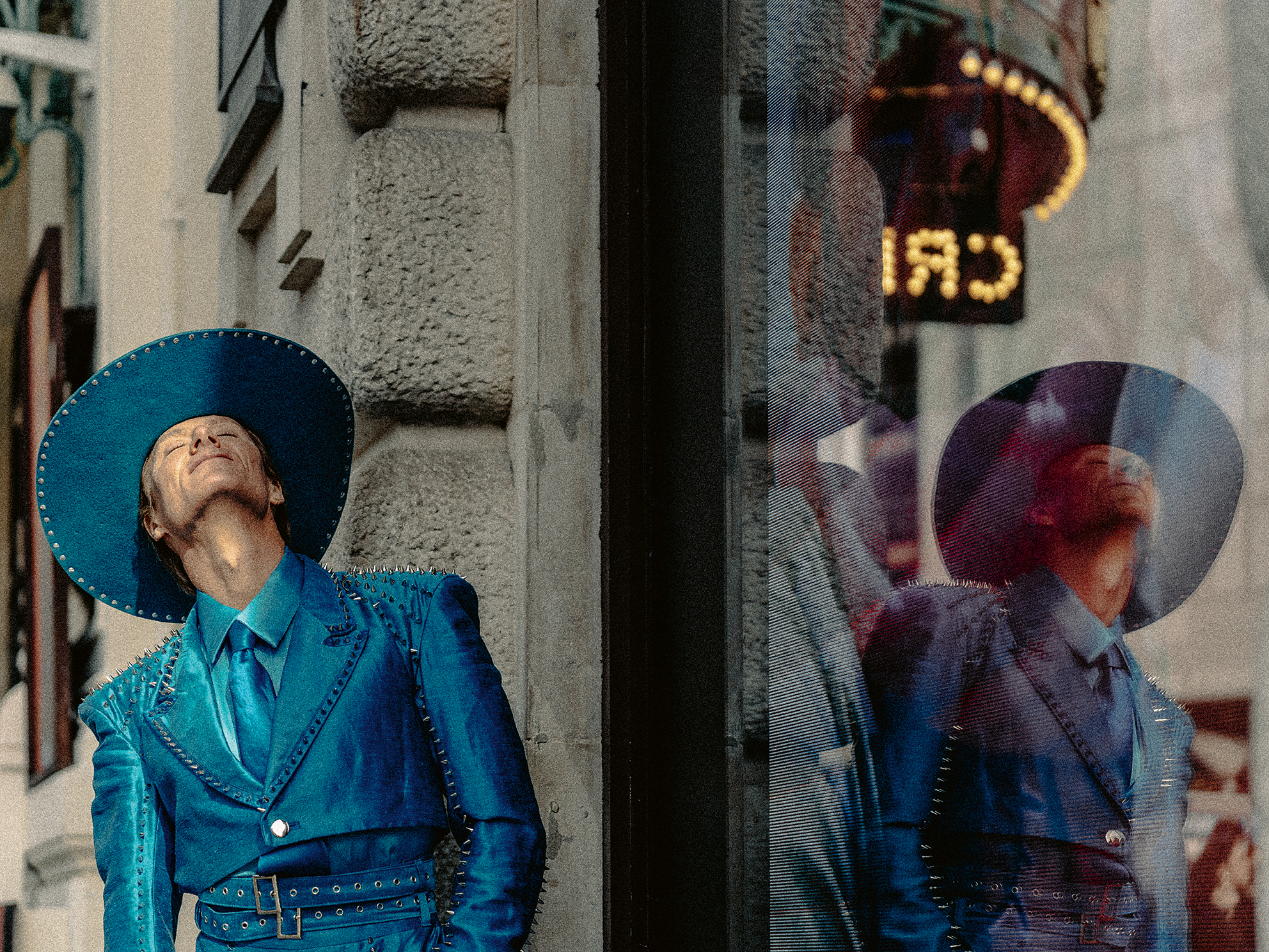 Person in studded blue suit and wide-brim hat leaning against a textured stone wall with their reflection in a nearby glass window