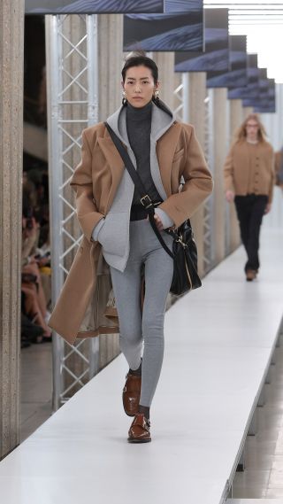 A model in Miu Miu Fall 2023 show wearing gray leggings, a gray turtleneck sweater, gray hoodie, tan top coat, and brown loafers.