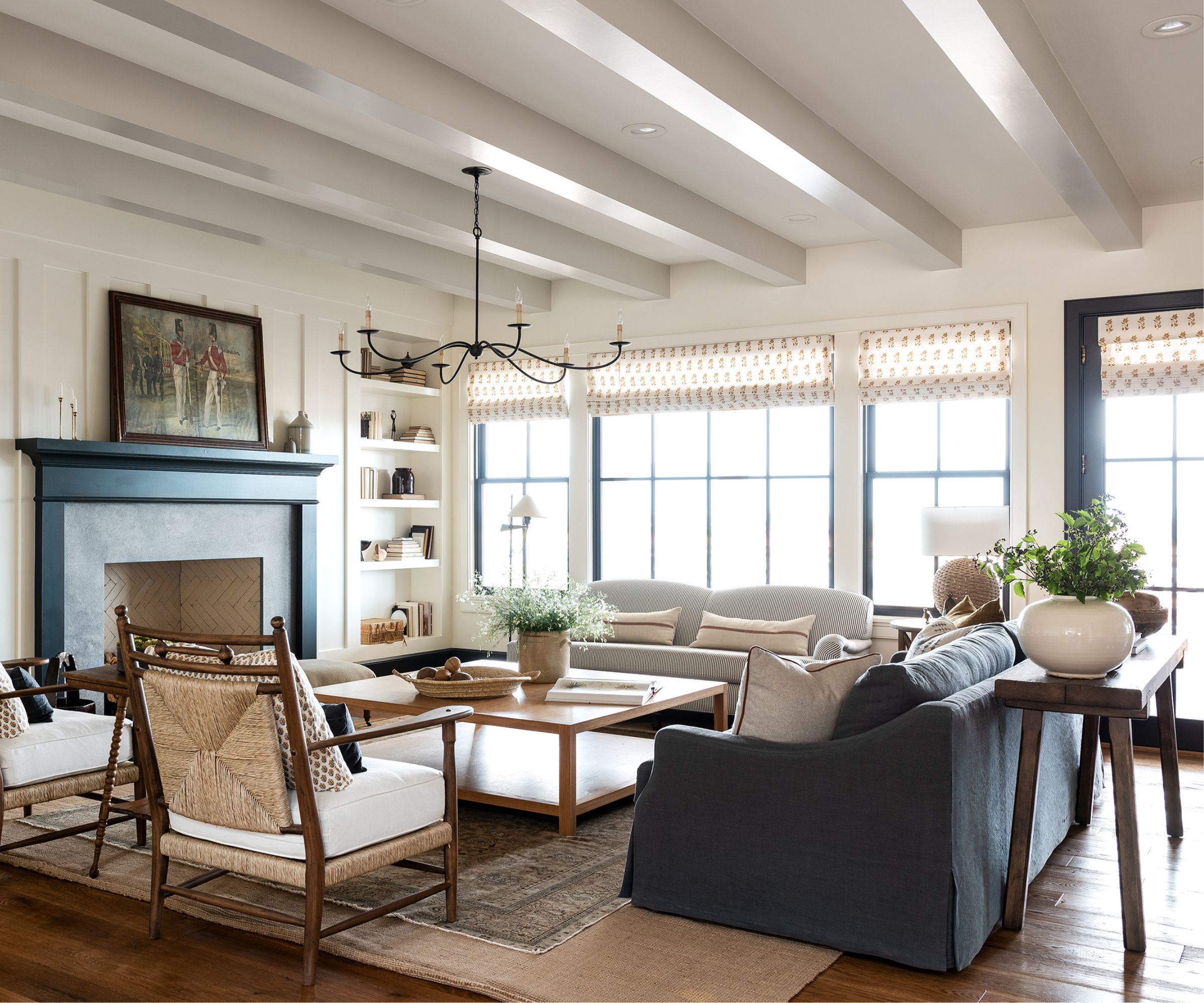 Modern farmhouse living room with white beamed ceiling, large windows with roman blinds, wooden floor, large fireplace, iron chandelier, slipcovered blue sofa, wooden coffee table, rattan chairs, and light blue loveseat