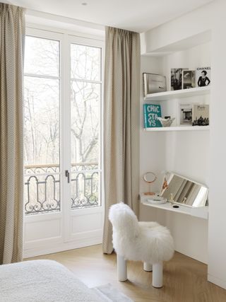 apartment bedroom with open shelving and sheepskin covered chair, mirror, dressing table/desk, books on shelves, neutral drapes, hardwood floor