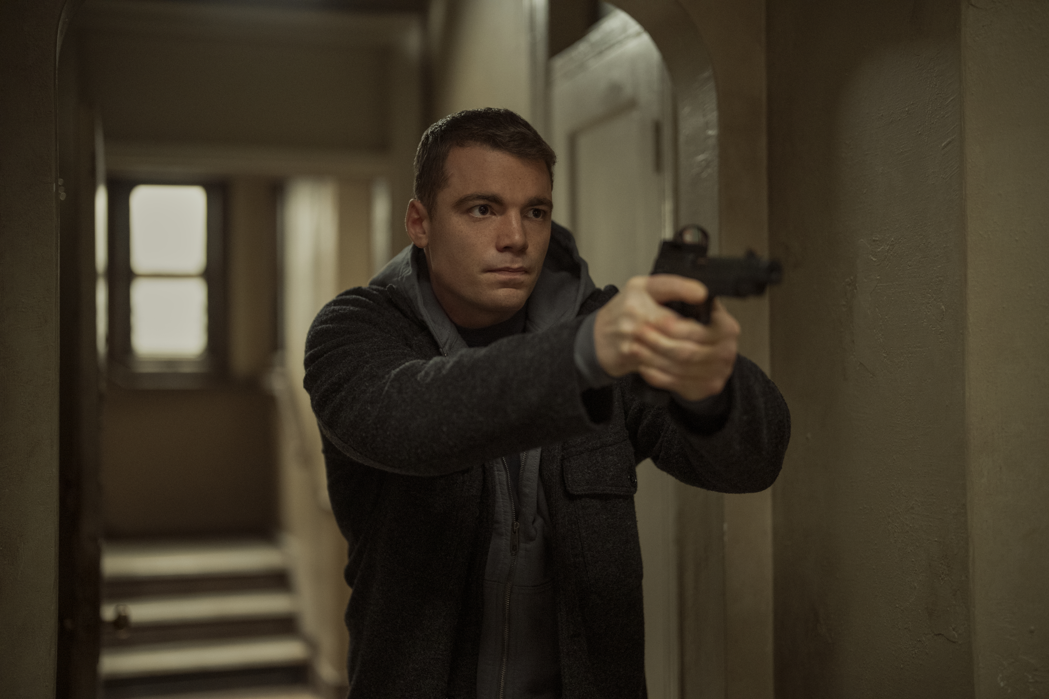 Gabriel Basso as Peter Sutherland pointing a gun walking down a dim hallway in a still from the night agent season 3
