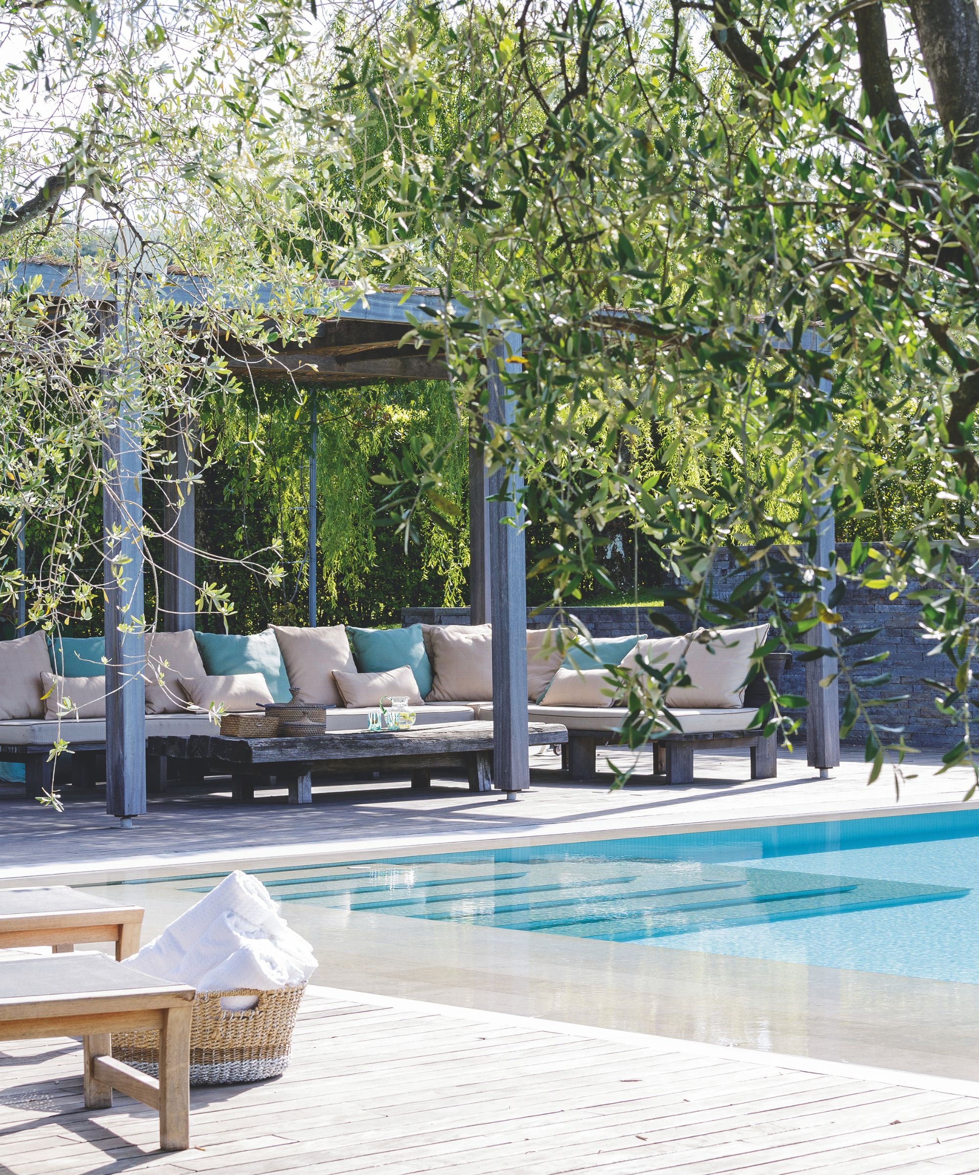 A leafy and sunny outdoor pool area with light washed decking, sun loungers with a wicker basket filled with white towels behind, and a large seating area with pink and blue cushions under a covered canopy beneath the trees.