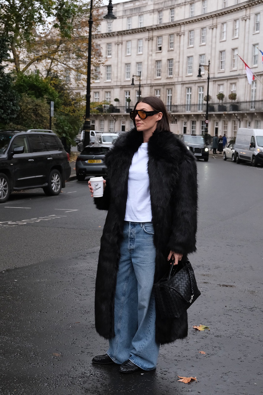A woman standing in the middle of the street in London wearing oversize two-tone aviator sunglasses with a long black fur coat layered over a white T-shirt, baggy wide-leg jeans, black ruched loafers, and a black Bottega Veneta Andiamo Bag.