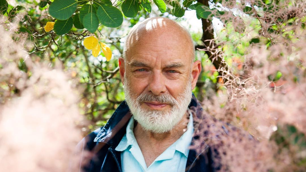 Brian Eno in a garden surrounded by leaves and flowers.