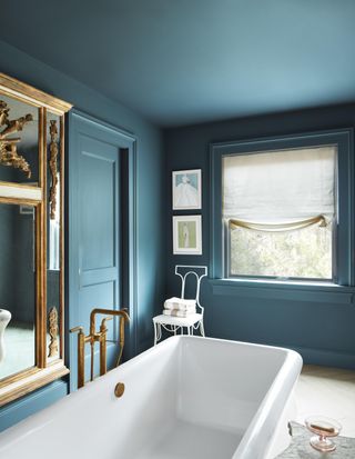 Corner of a blue color-drenched bathroom with a white tub and ornate gold mirror in view