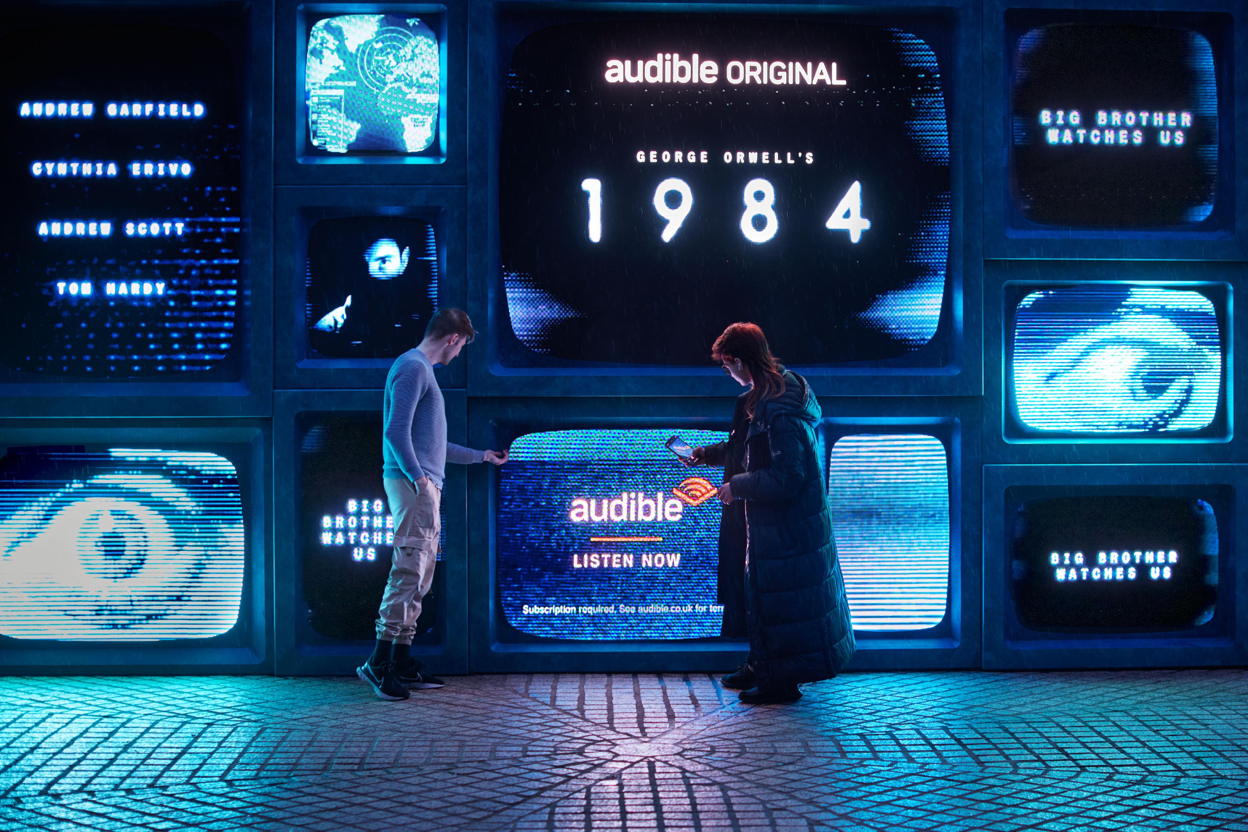 Audible 1984 on big screens with two people walking past looking at the screens