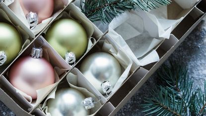 A box of pastel colored baubles with cardboard dividers between them. Pieces of fir tree cut up and laying around the box. 