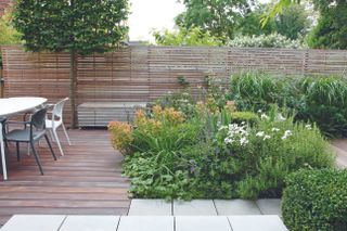 Patio in a decked garden with slatted wooden fence and modern white table and chairs.
