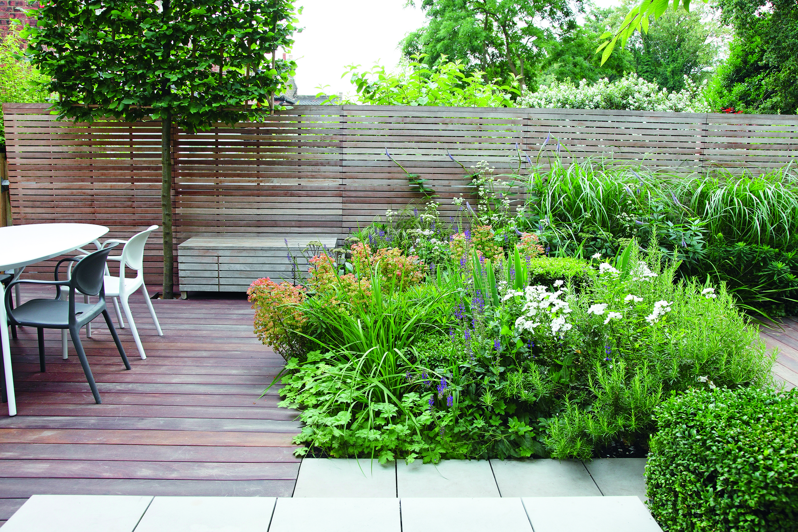 Patio in a decked garden with slatted wooden fence and modern white table and chairs.