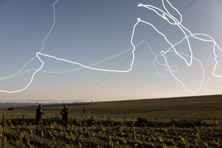 Daytime image, white squiggly lines representing the trail of the aeroglyphic when in flight, two men holding on to a rope cord, vineyard, grassed landscape, clear pale blue sky