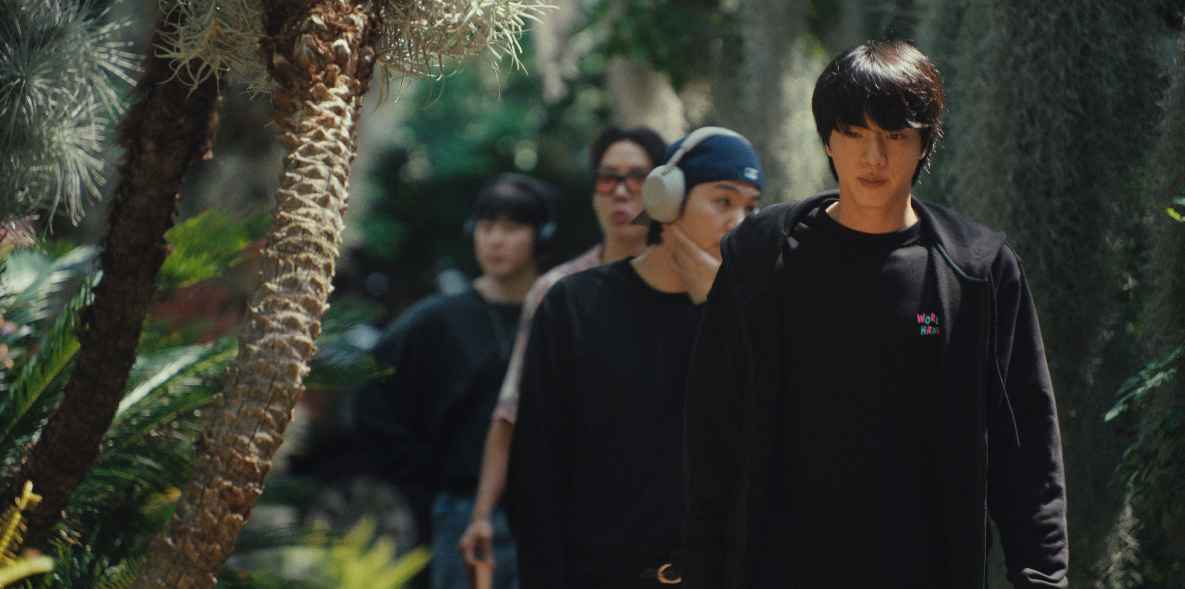 Some BTS members (l-r: Jimin, j-hope, Suga, and Jin) walk down a garden path lined with palm trees, in the Netflix documentary 'BTS: THE RETURN.'