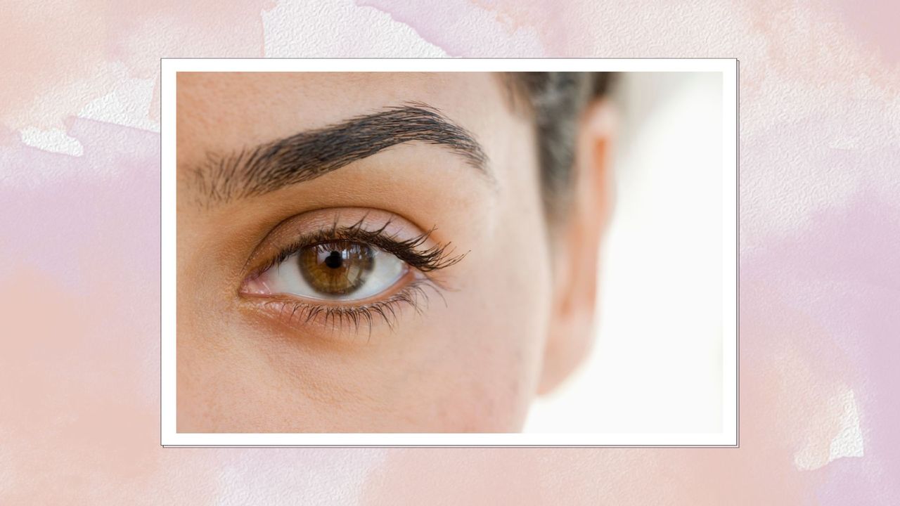 A close up of a woman&#039;s eye with full, well groomed eyebrows on a pink watercolour background