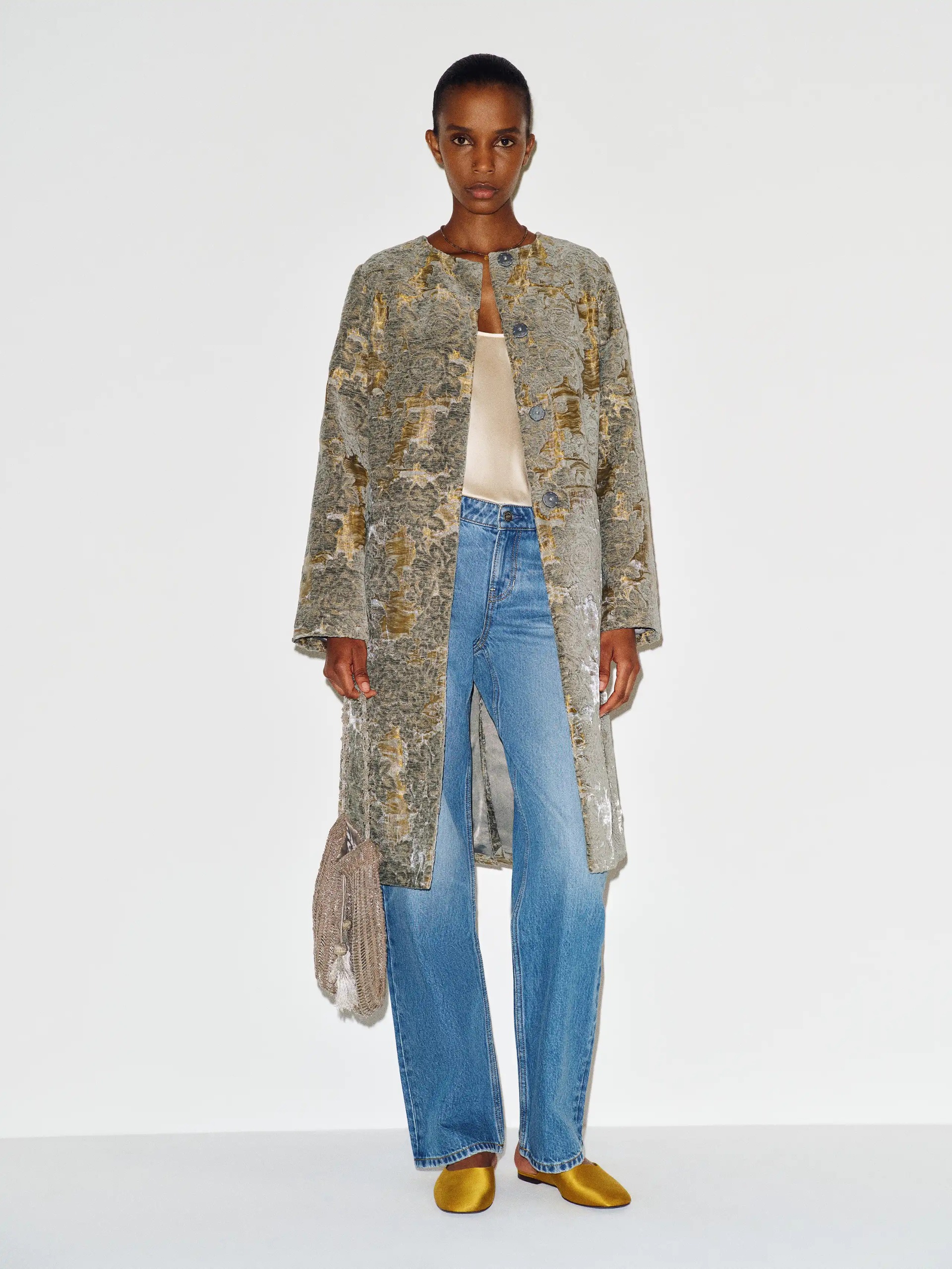 An image of a model wearing a brocade coat in silver and gold with blue jeans and yellow satin mules