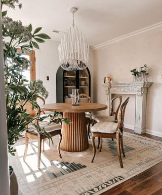 A dining room with a wooden round table and wooden chairs with cushions around it, a large chandelier above it and a white patterned rug below it, and a large leafy plant to the left