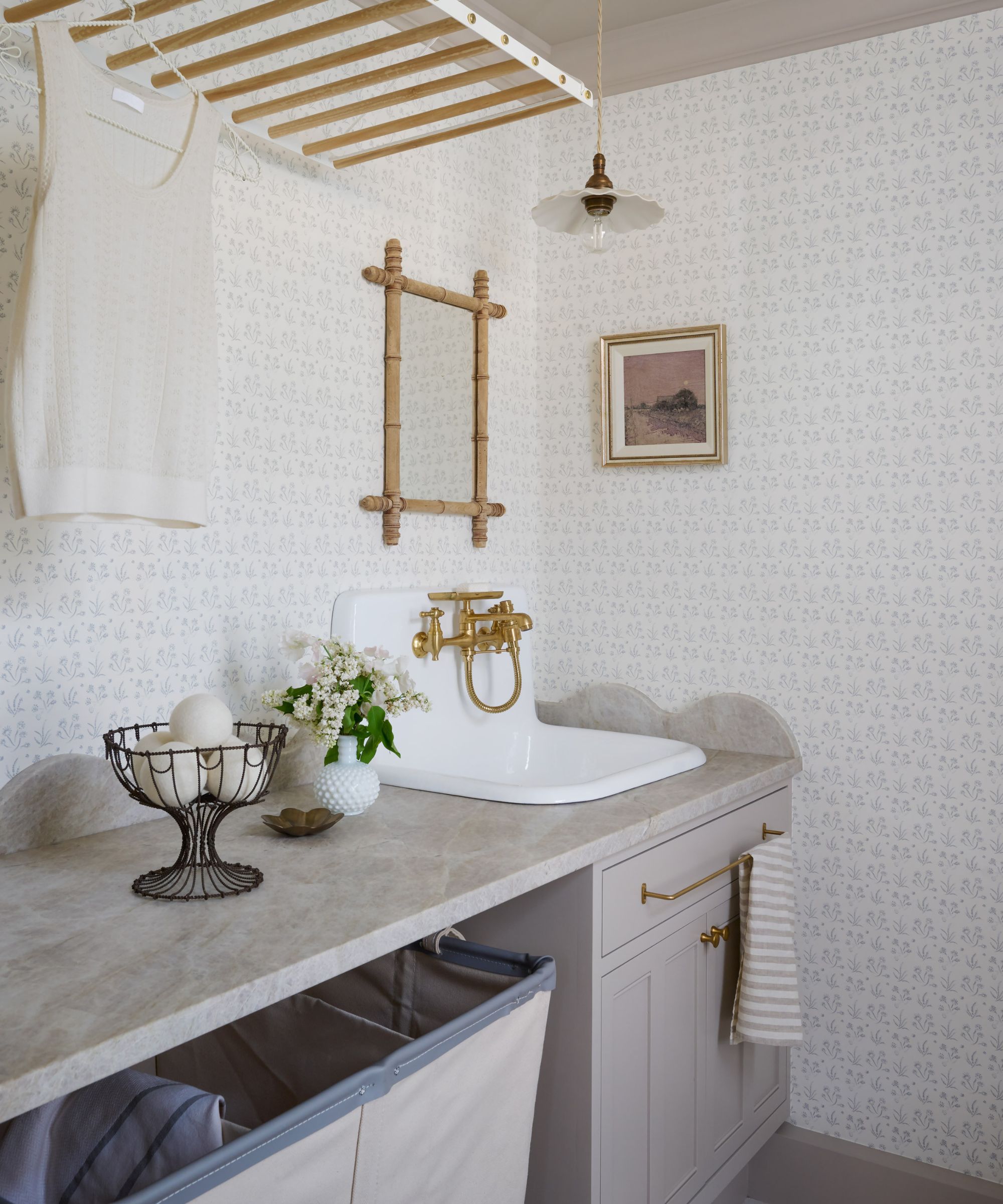 Laundry room with wallpapered walls, Elephant's Breath by Farrow &amp;amp; Ball painted cabinetry, a ceramic sink with a gold faucet, a laundry bin underneath, and a wood hanging rack above.