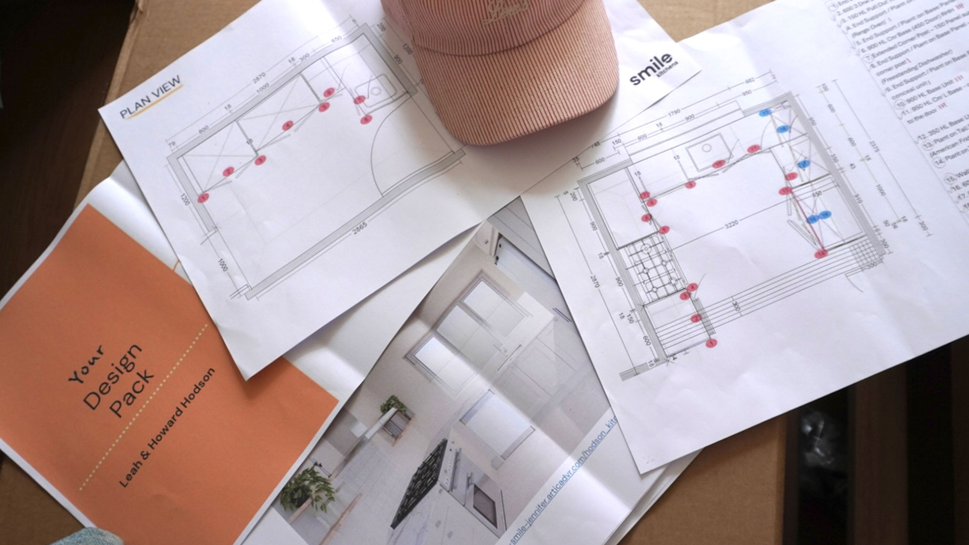 Paper plans for a kitchen laid out over a table, with a pink baseball cap resting on top