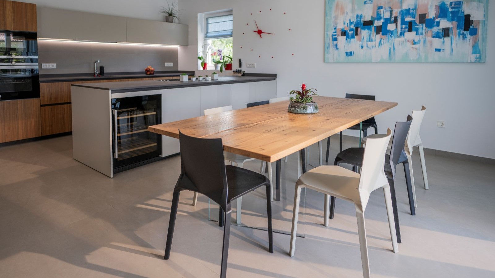 A cement floor with wooden kitchen units and a white kitchen island with a blue canvas painting on the wall. A table and chairs is in the foreground