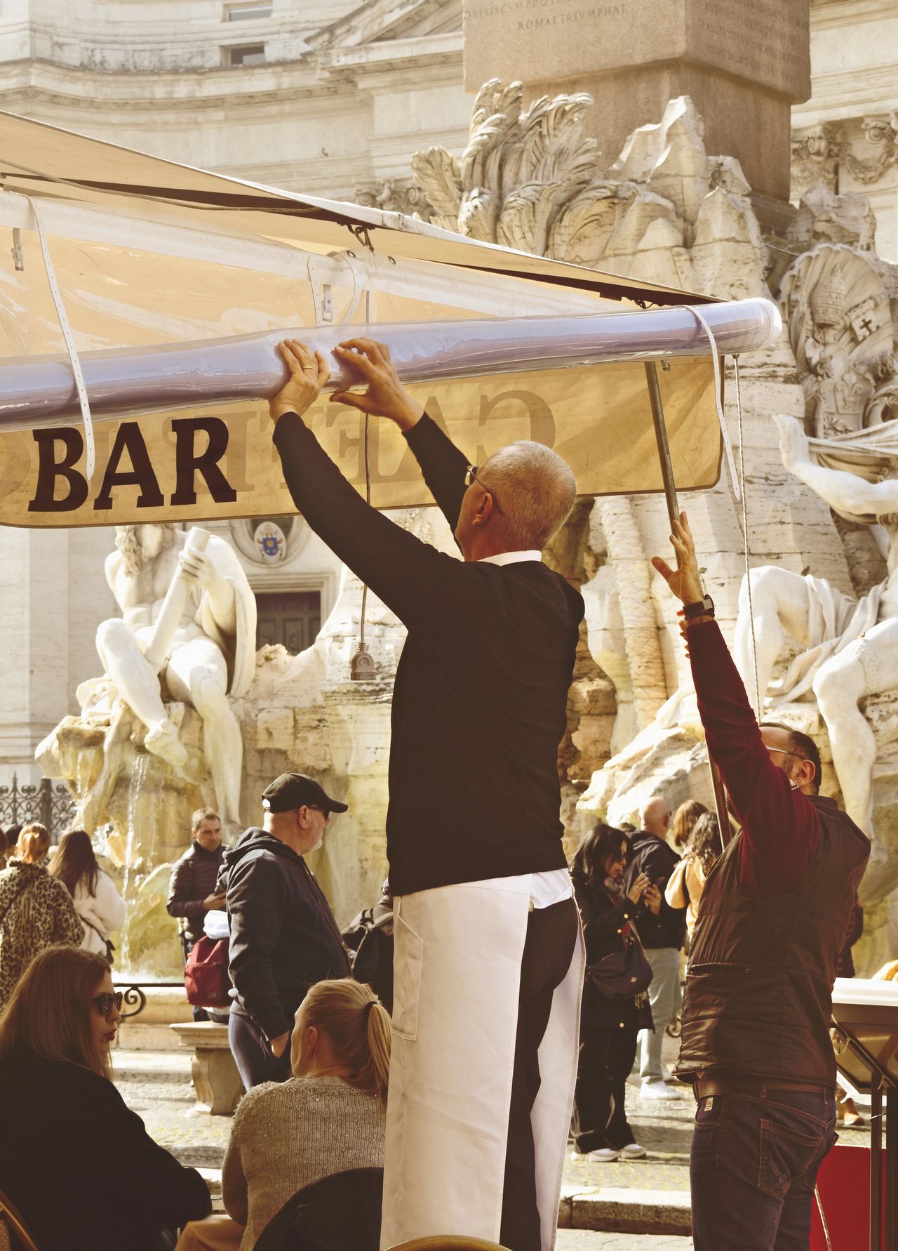 Waiter folding up a restaurant awning infront of a big marble sculpture in Rome