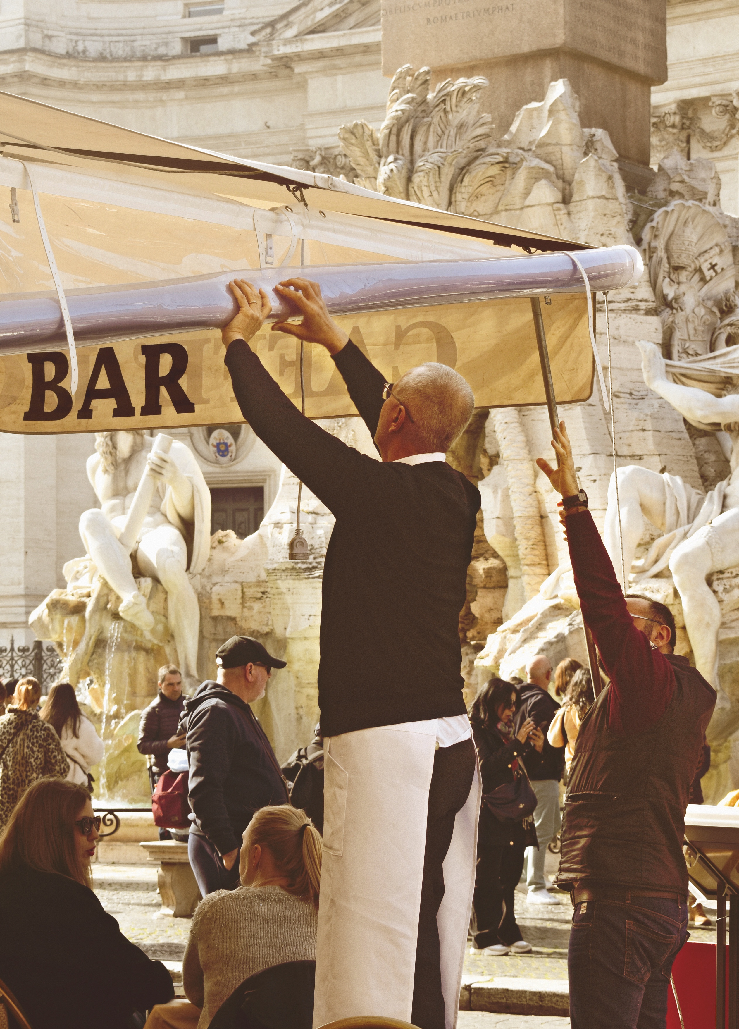 Waiter folding up a restaurant awning infront of a big marble sculpture in Rome