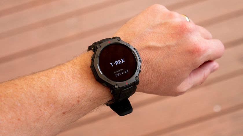 A photo of the Amazfit T-Rex 3 Pro on a man&#039;s wrist above a wooden deck, showing the boot-up screen with &quot;T-REX Powered by Zepp OS&quot; visible.